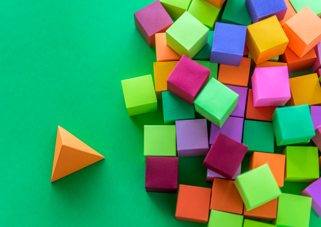 Lots of different coloured cubes on top of each other to the right on a green background with a single orange pyramid standing out next to them to the left on the green background